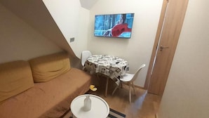 Un petit salon avec un canapé, une table ronde recouverte d’une nappe à motifs de journaux et un téléviseur fixé au mur.