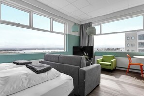 Una habitación de hotel moderna con una cama grande, un sofá gris, un sillón verde y una pequeña mesa con una silla naranja.