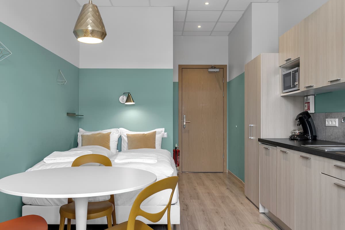 Une pièce de taille modeste comprenant un lit, une table ronde avec des chaises, un coin cuisine et une porte donnant sur une autre pièce.