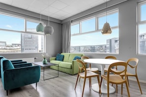 A modern living room with a green sofa, teal armchairs, and wooden chairs around a white table. Large windows offer a city view. - Icelandic Apartments (Kópavogur)