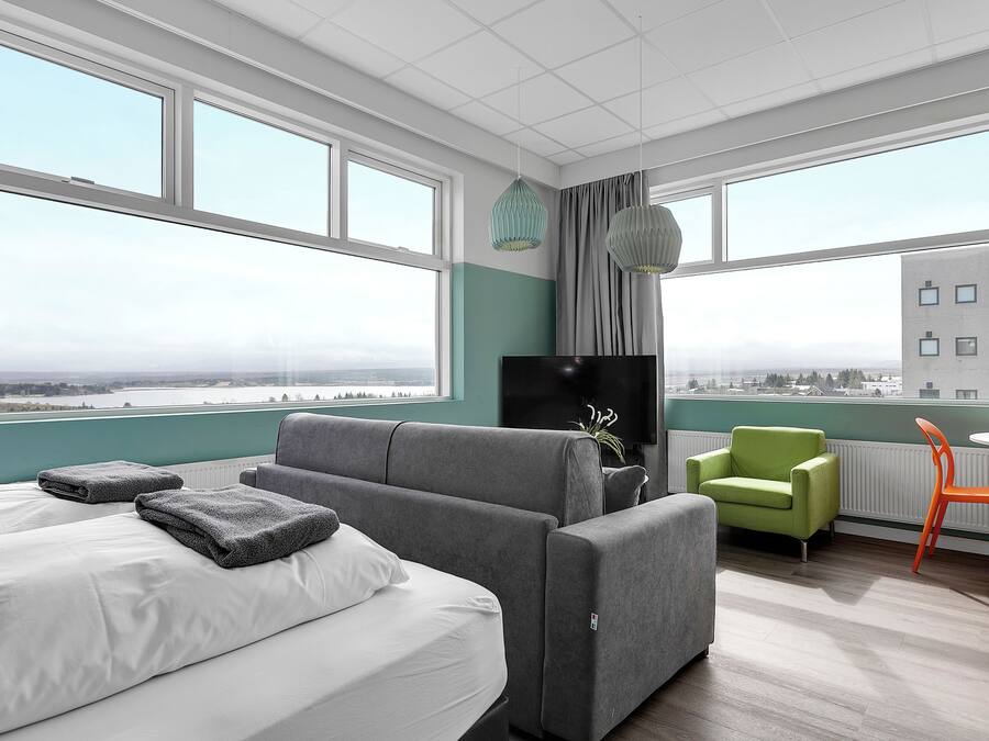 A modern hotel room with a large bed, a grey sofa, a green armchair, and a small table with an orange chair.