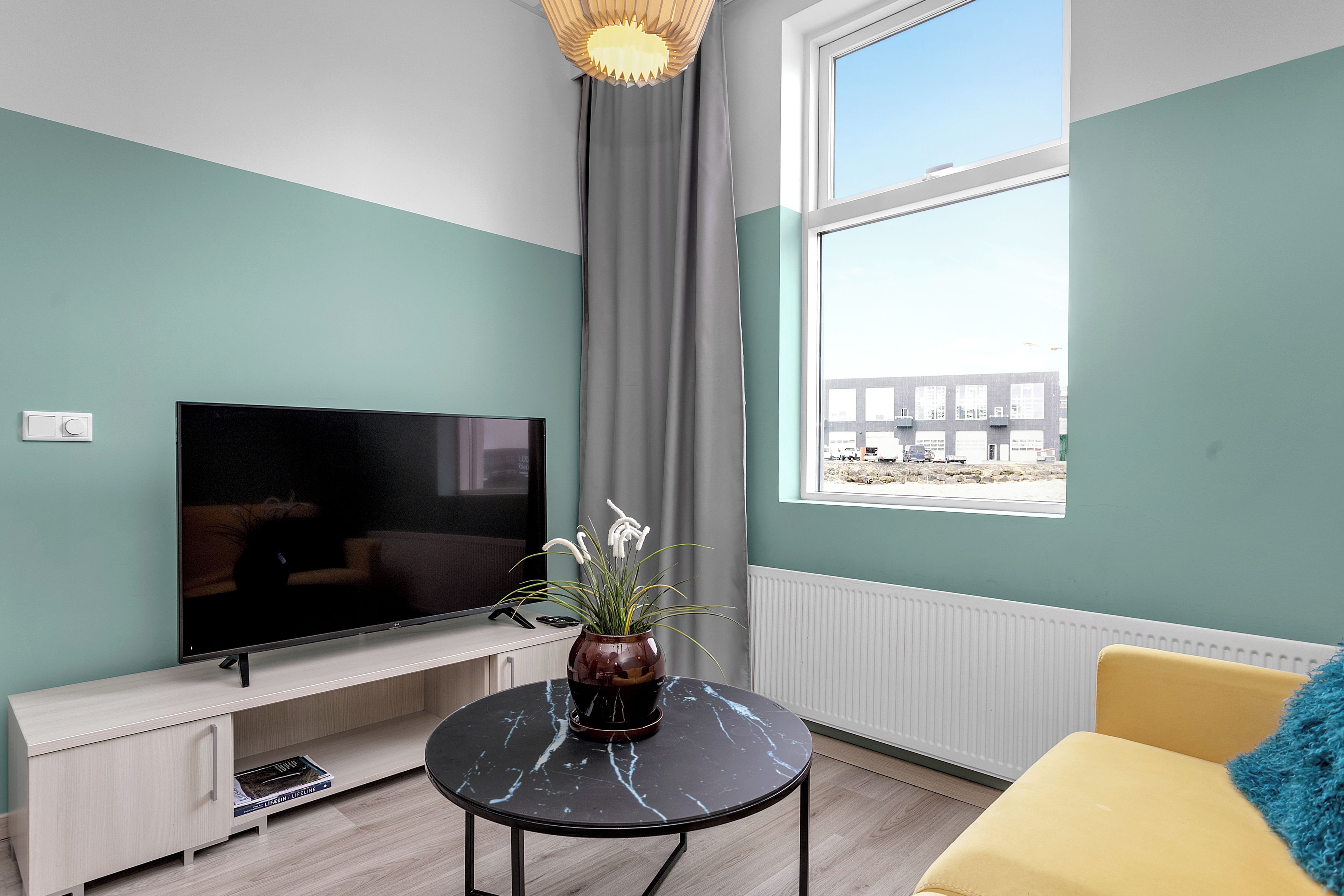 Una sala de estar moderna con una televisión de pantalla plana, una mesita con un florero, un sofá amarillo y una ventana con vistas a edificios.