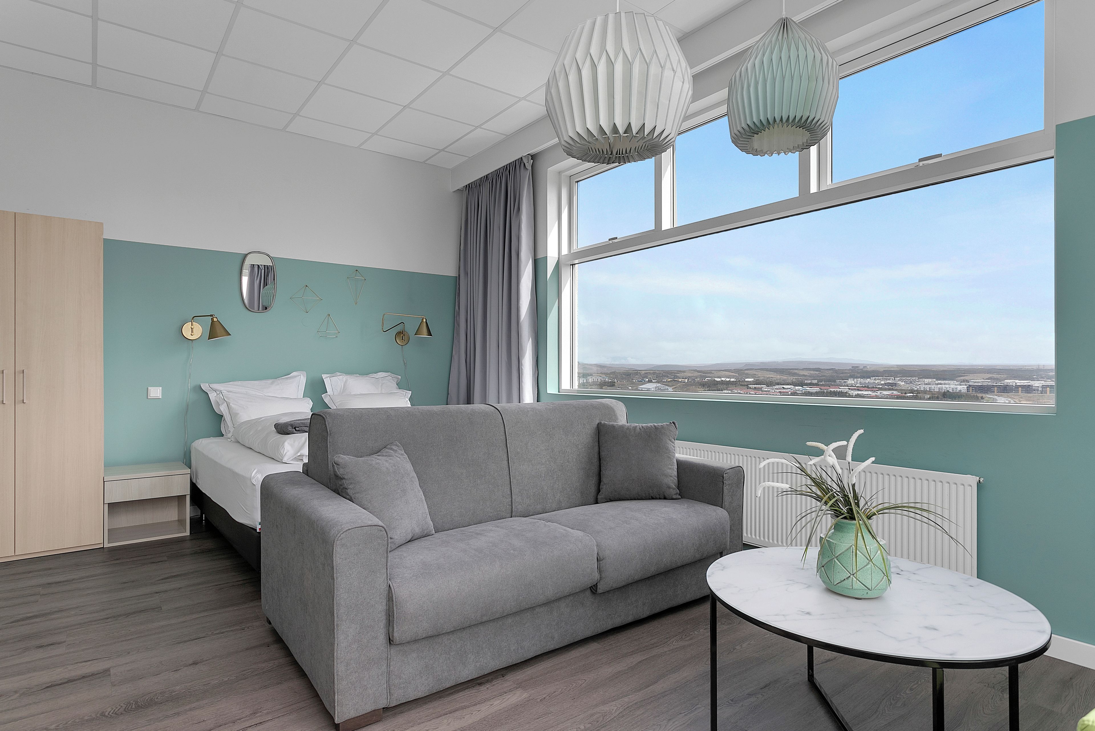 Une chambre moderne avec un canapé gris, une petite table ronde ornée d’un vase, un lit avec une literie blanche et une vue sur la ville grâce à de grandes fenêtres.