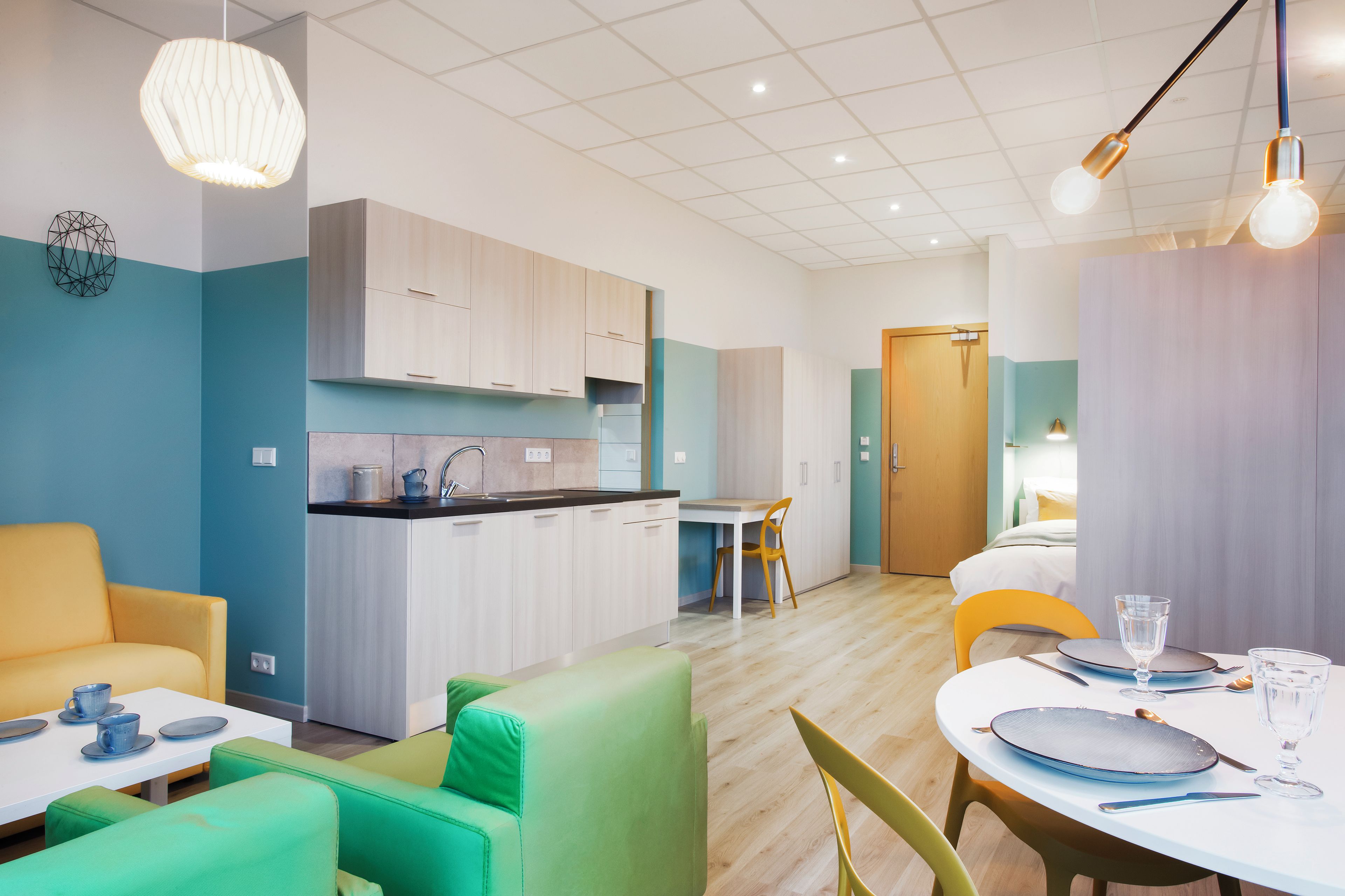 Une cuisine moderne avec des meubles blancs, un coin repas avec une table dressée pour deux personnes, et un salon avec des canapés verts.