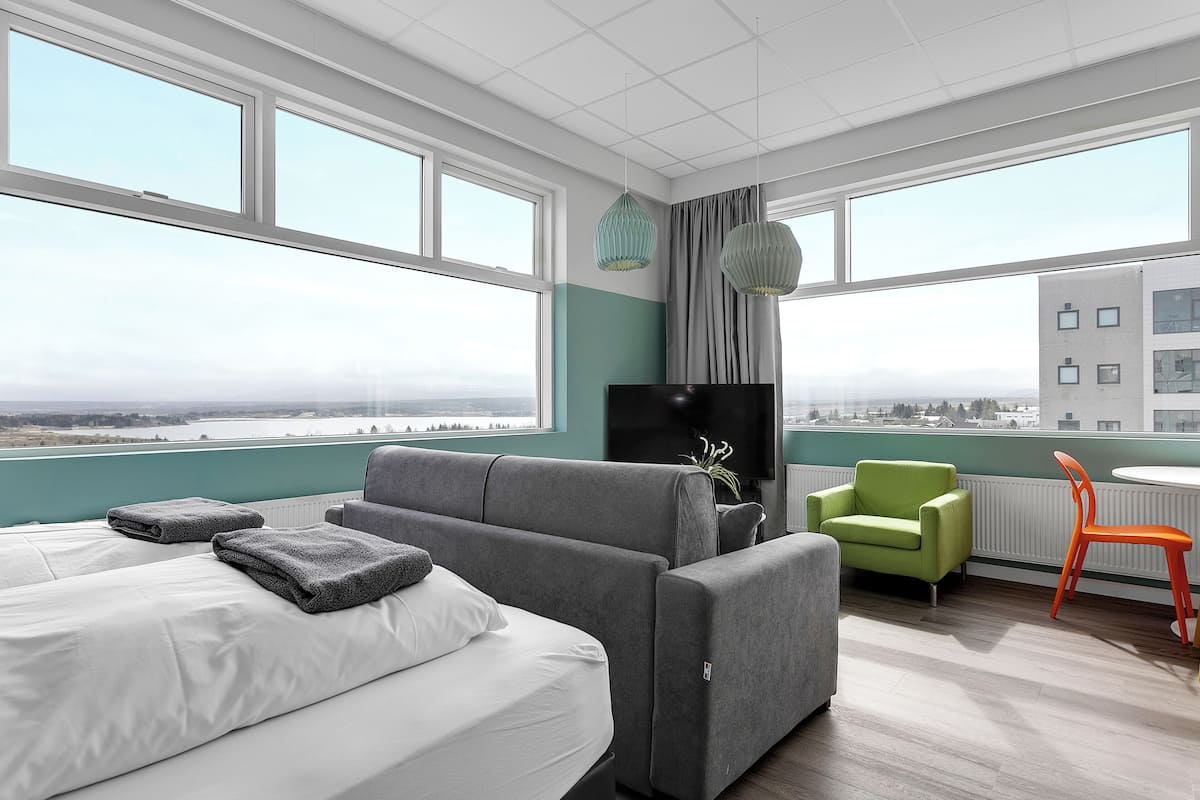 Une chambre d’hôtel moderne avec un grand lit, un canapé gris, un fauteuil vert et une petite table avec une chaise orange.