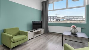 Ett modernt rum med turkosa väggar, en grön soffa, en tv, ett element och utsikt över en byggarbetsplats.