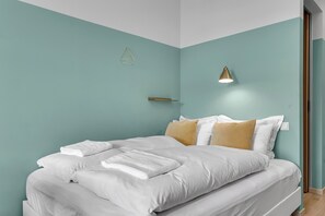 A neatly made bed with white linens and a single brown pillow, a gold lamp on the bedside table, and a geometric wall decoration. - Icelandic Apartments (Kópavogur)