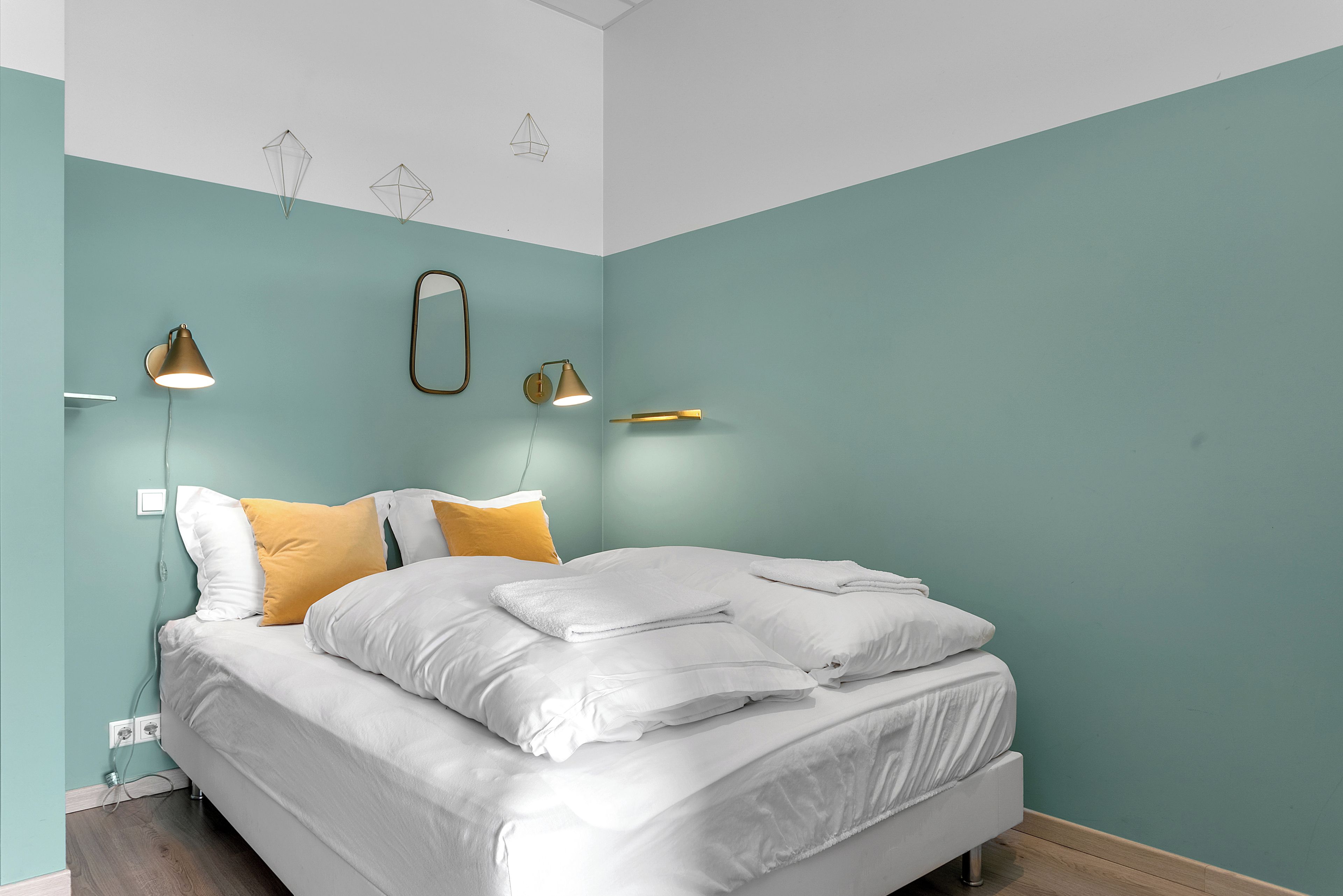 Cama bem arrumada com almofadas brancas e amarelas, abajur e espelho fixado na parede.