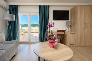A hotel room with a sofa, a round table with a vase of flowers, a TV on the wall, and a sliding door leading to a balcony. - Hotel Porat (Neum)