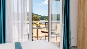 A bedroom with a large bed, a sliding glass door leading to a balcony with a table and chairs, and a view of a lake and hills. - Hotel Porat (Neum)