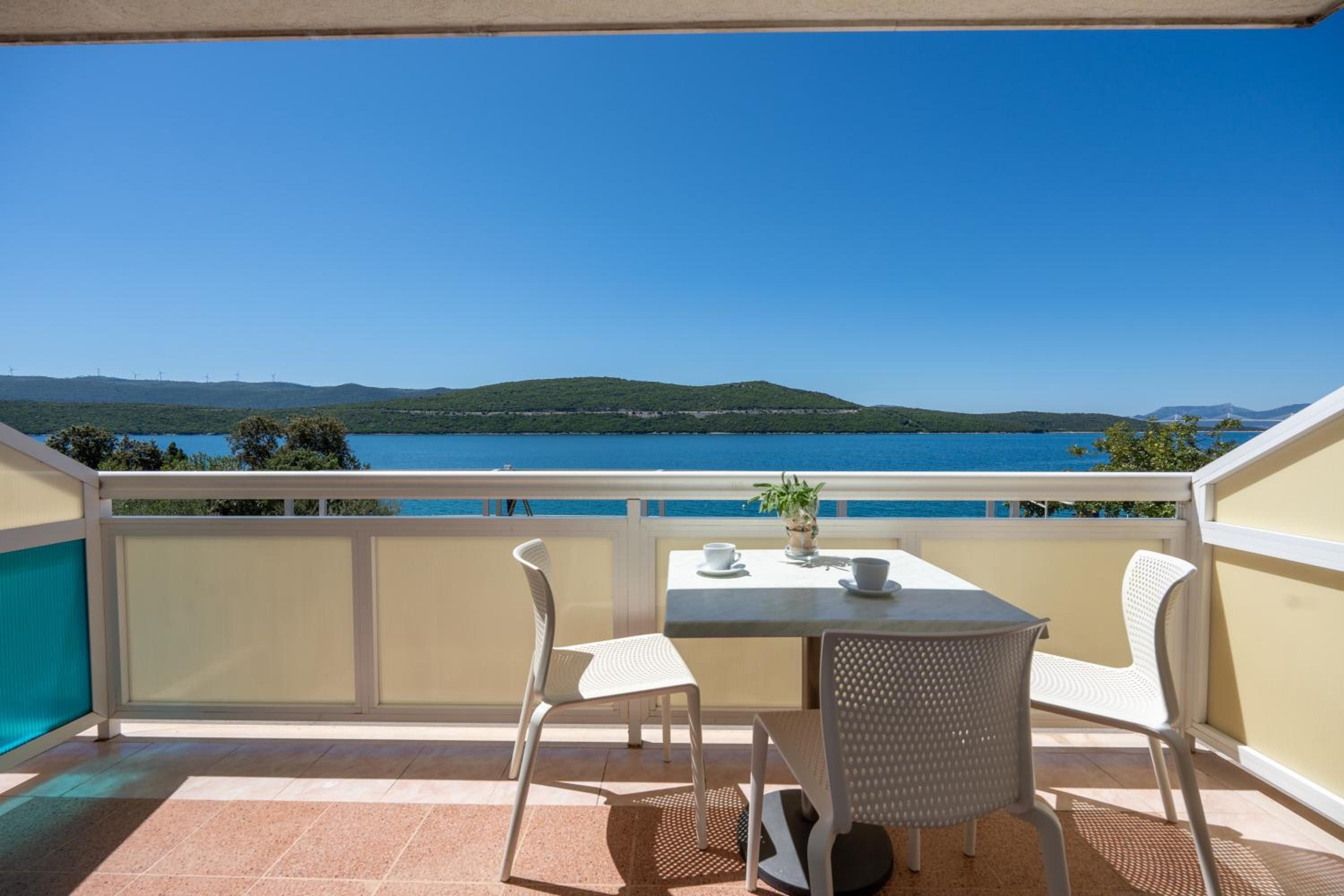 Un balcón con mesa y sillas, con vista a una masa de agua y colinas al lejano.