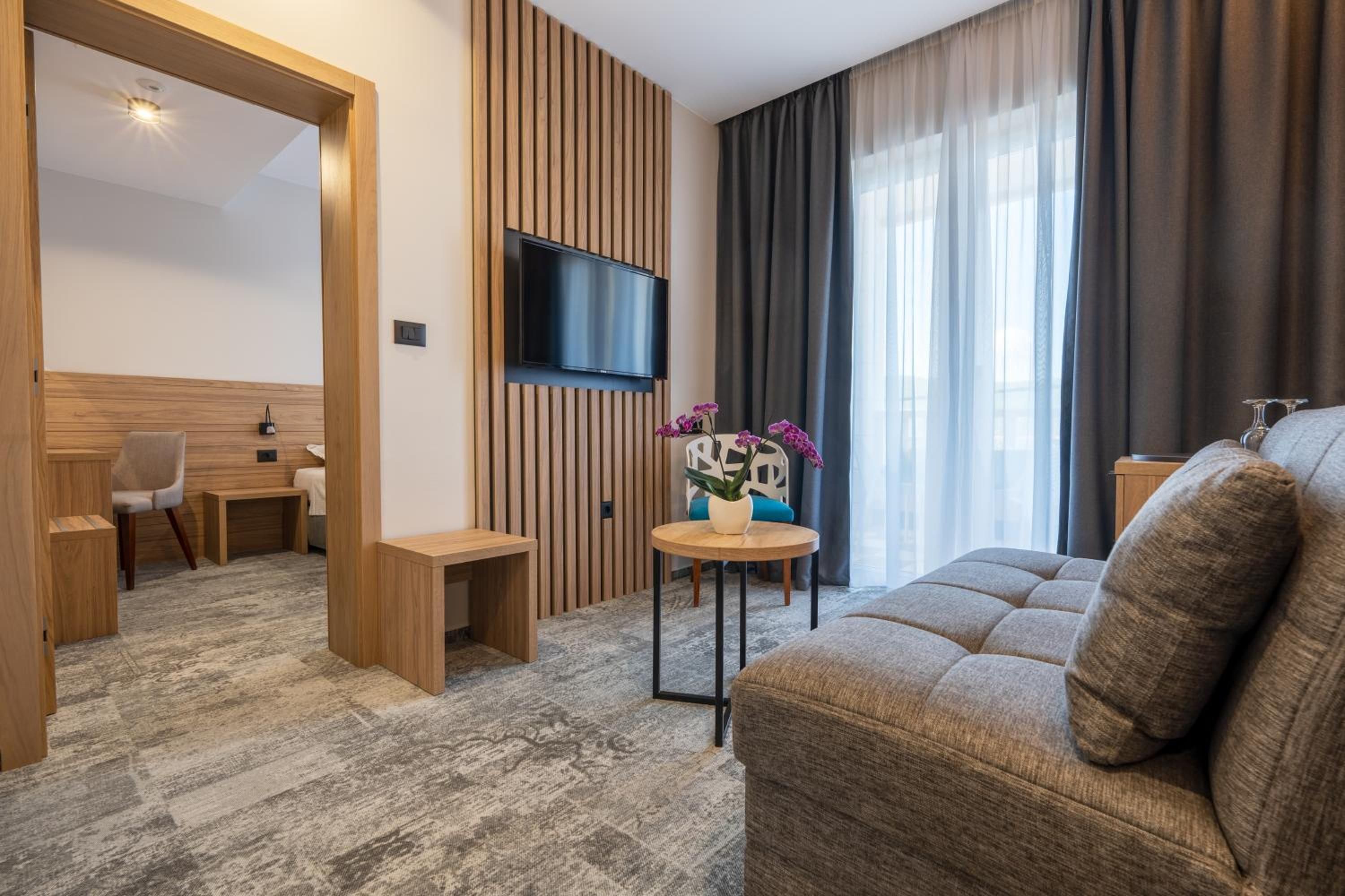 A modern hotel room with a flat-screen TV mounted on a wooden panel wall, a sofa, a small round table with a potted plant, and a chair.