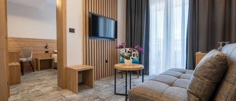 A modern hotel room with a flat-screen TV mounted on a wooden panel wall, a sofa, a small round table with a potted plant, and a chair.