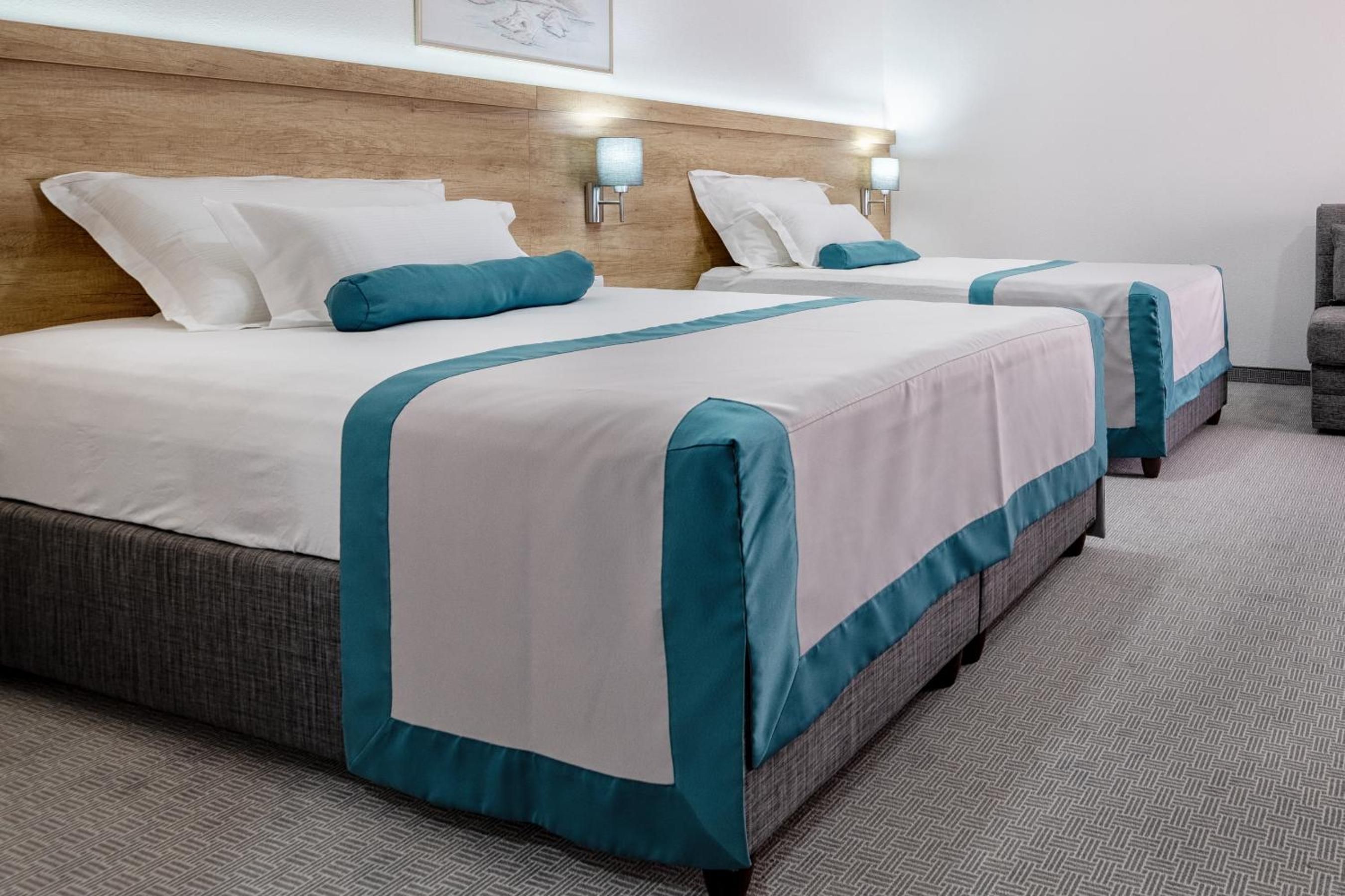 A hotel room with two beds, each with white bedding and teal accents, a wooden headboard, wall-mounted lamps, and a herringbone-patterned carpet.