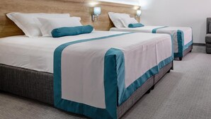 A hotel room with two beds, each with white bedding and teal accents, a wooden headboard, wall-mounted lamps, and a herringbone-patterned carpet. - Hotel Porat (Neum)