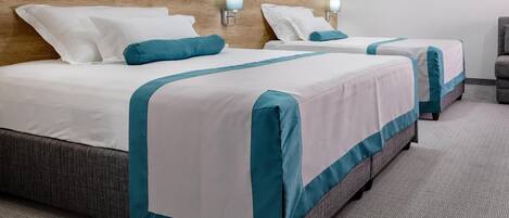 A hotel room with two beds, each with white bedding and teal accents, a wooden headboard, wall-mounted lamps, and a herringbone-patterned carpet.