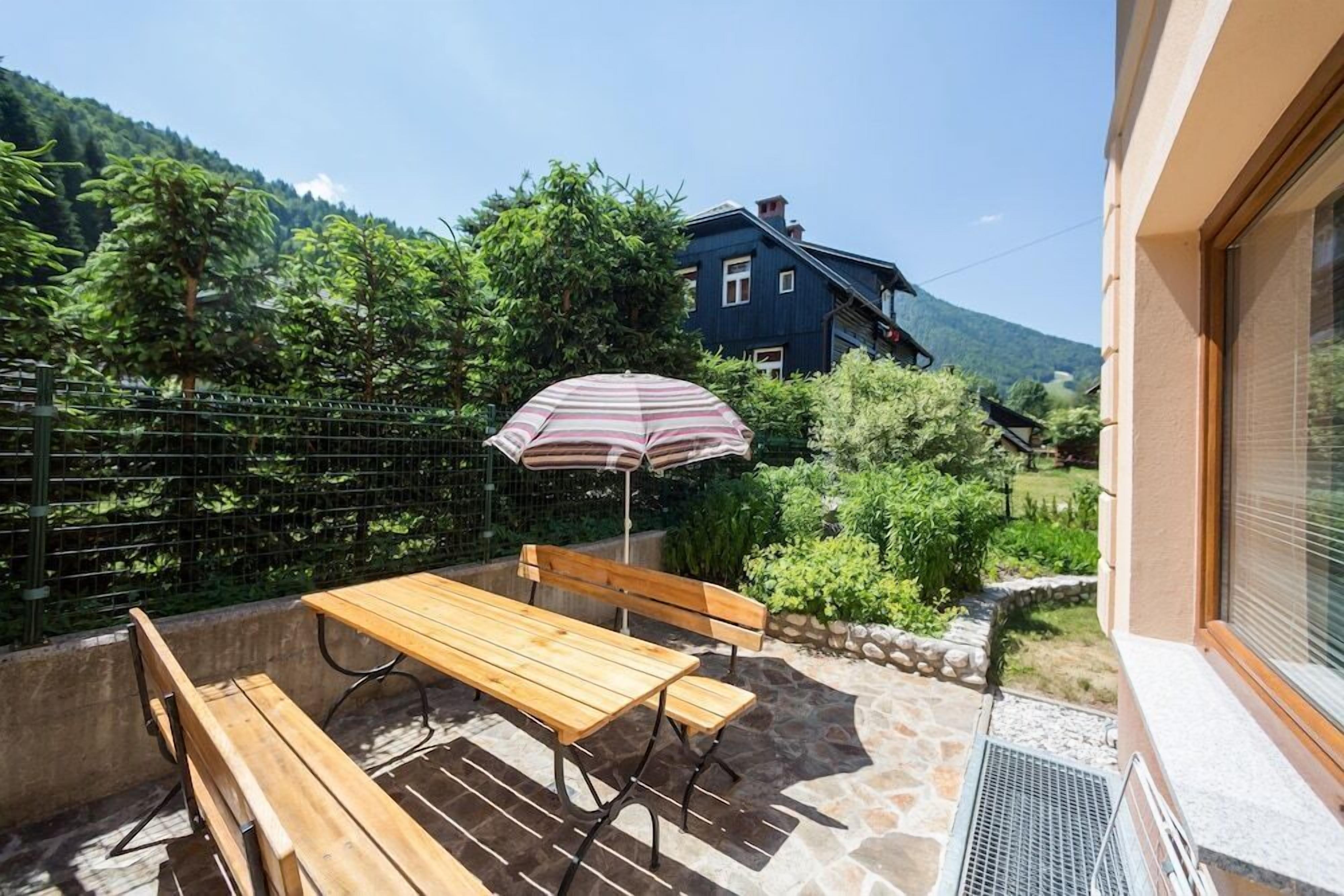 Um pátio com mesa e banco de madeira, um guarda-sol e vista para uma casa e montanhas.