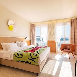 Hotellihuone, jossa on sänky, tuoli, ikkuna verhoilla ja näkymä kaupunkiin.
