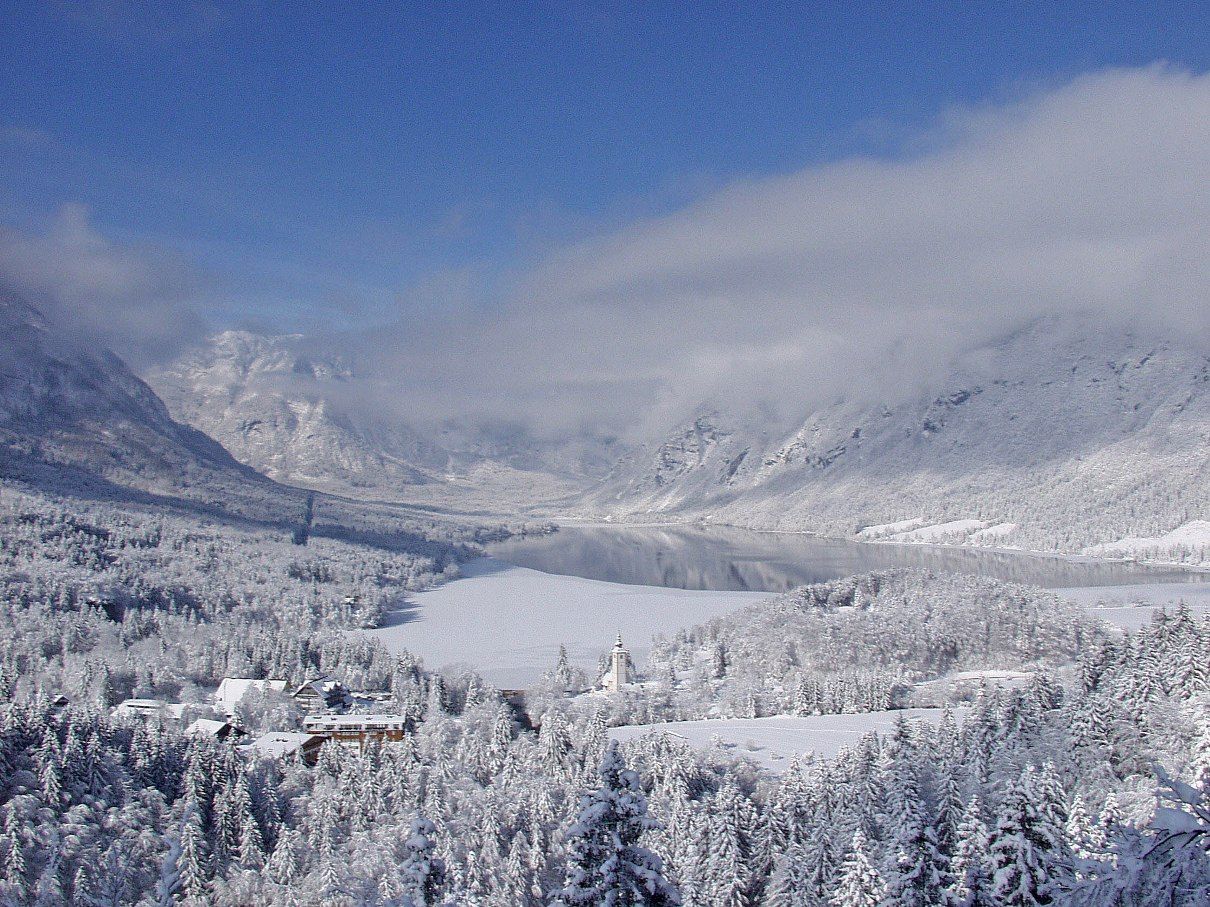 A snow-covered landscape with a lake, mountains, and a building with a tower.