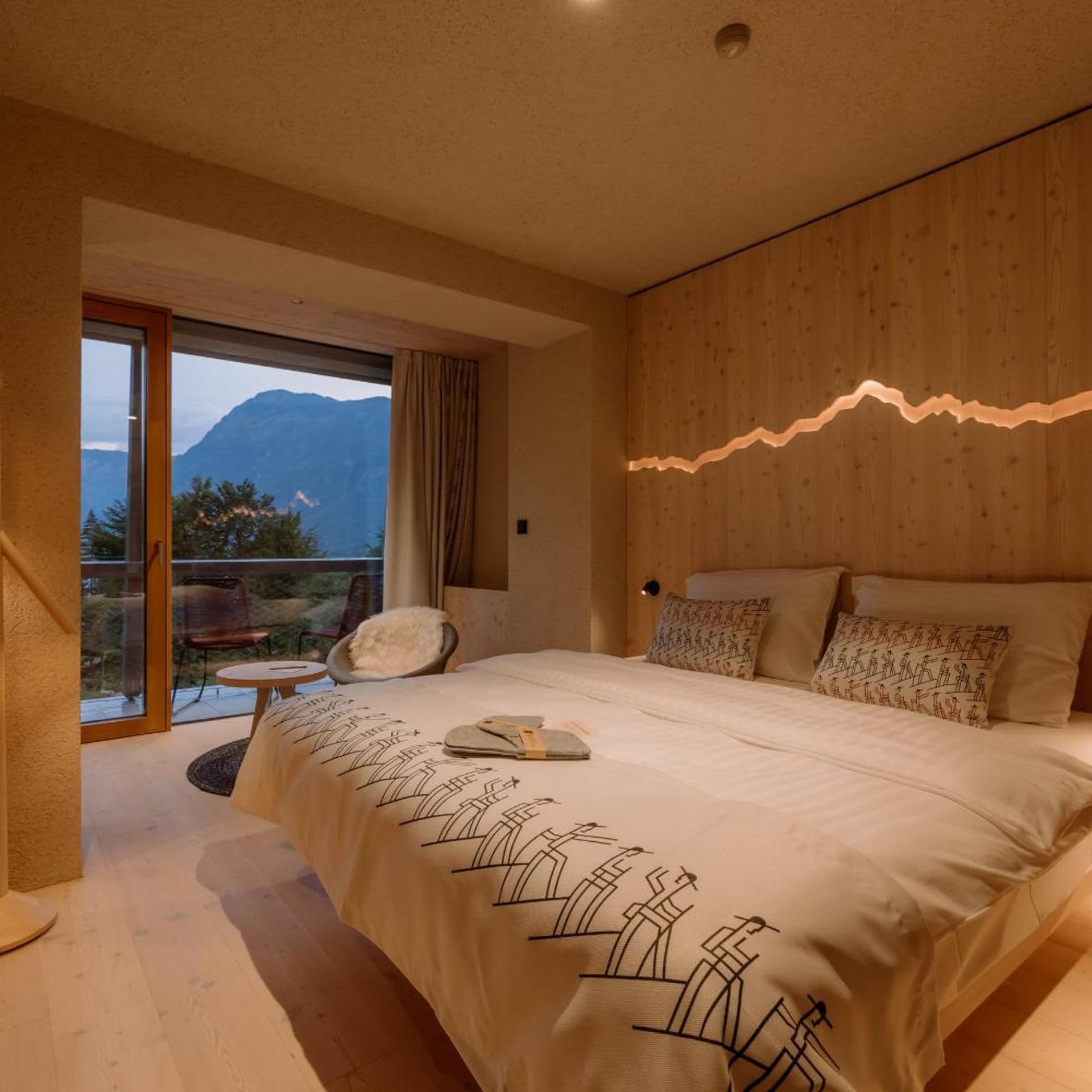 A bedroom with a bed, a chair, a table, and a view of mountains through a glass door.