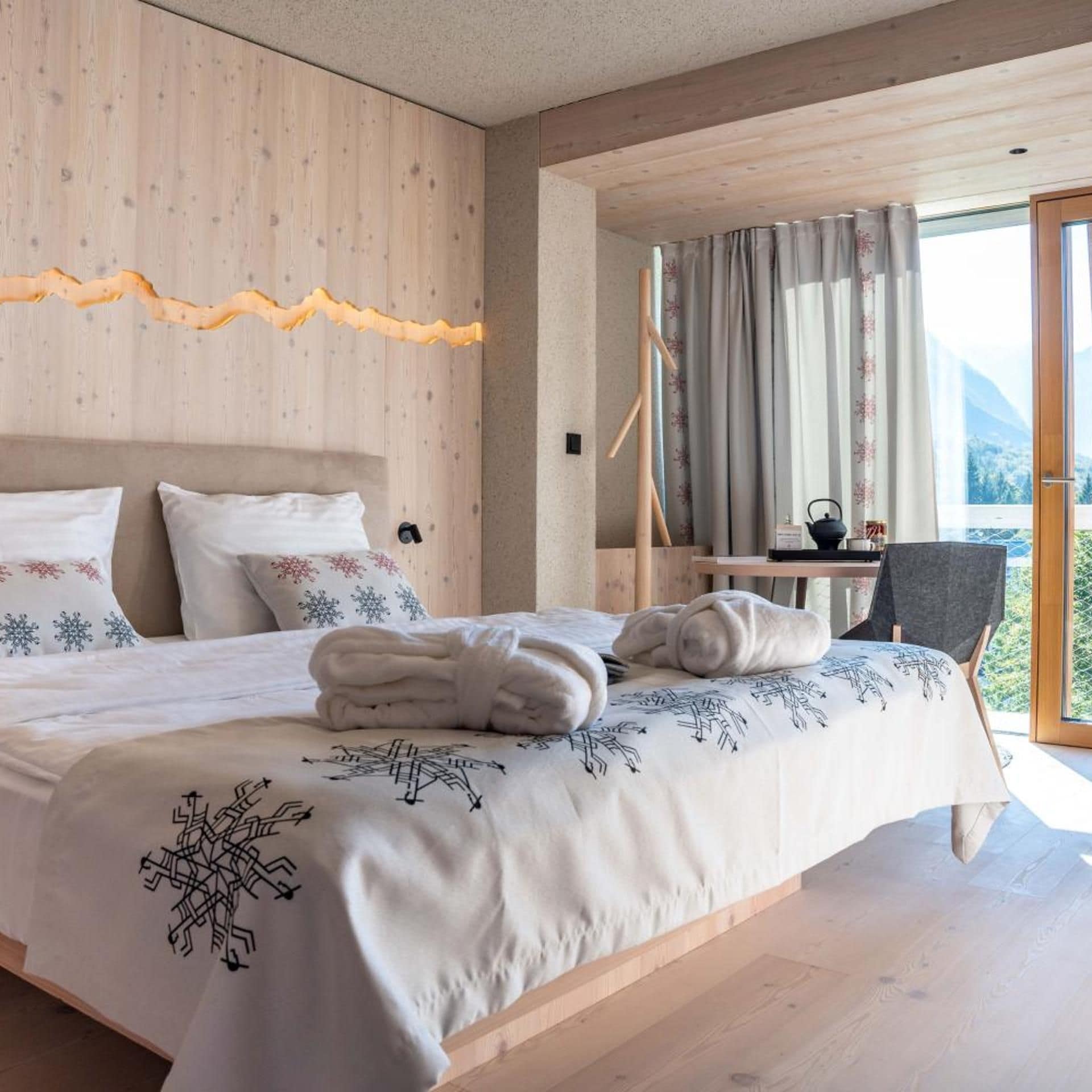 A modern bedroom with a wooden headboard, a bed with a snowflake-patterned bedspread, a bedside table with a kettle, and a view of the outdoors.