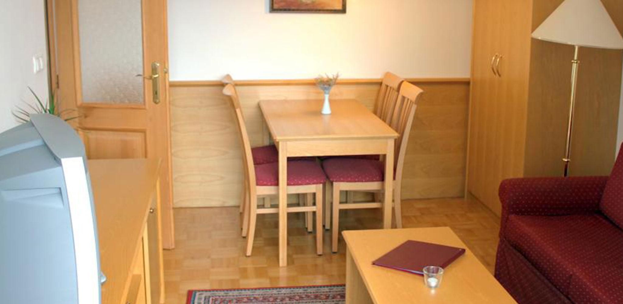 Une pièce de taille modeste, avec une table à manger en bois, deux chaises, une petite table sur laquelle se trouvent un livre et un verre, un canapé rouge, une lampe et un tableau accroché au mur.