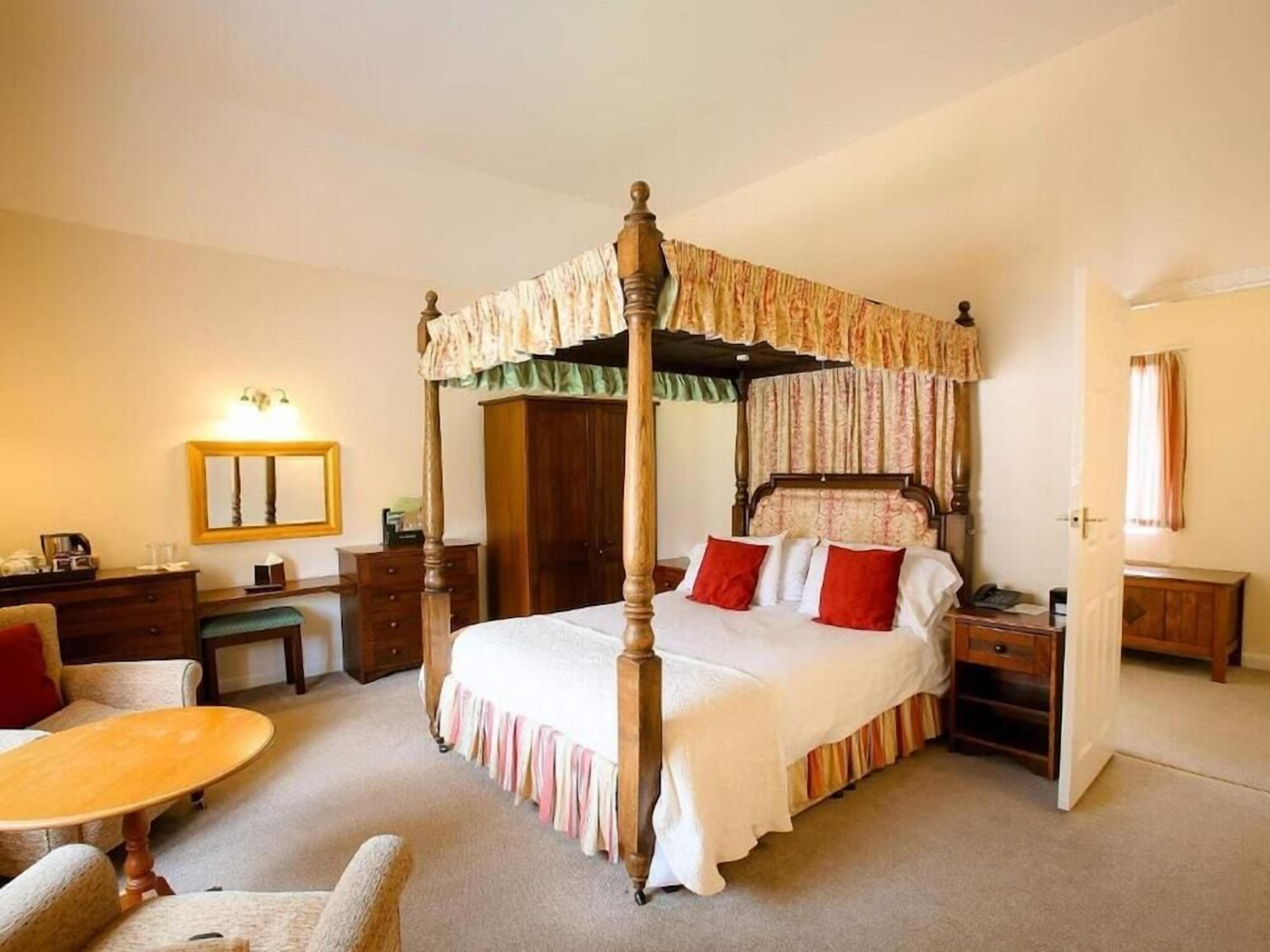 A four-poster canopy bed with a wooden frame and a green and white canopy, a bedside table with a lamp, a chair, a small round table, and a mirror on the wall.