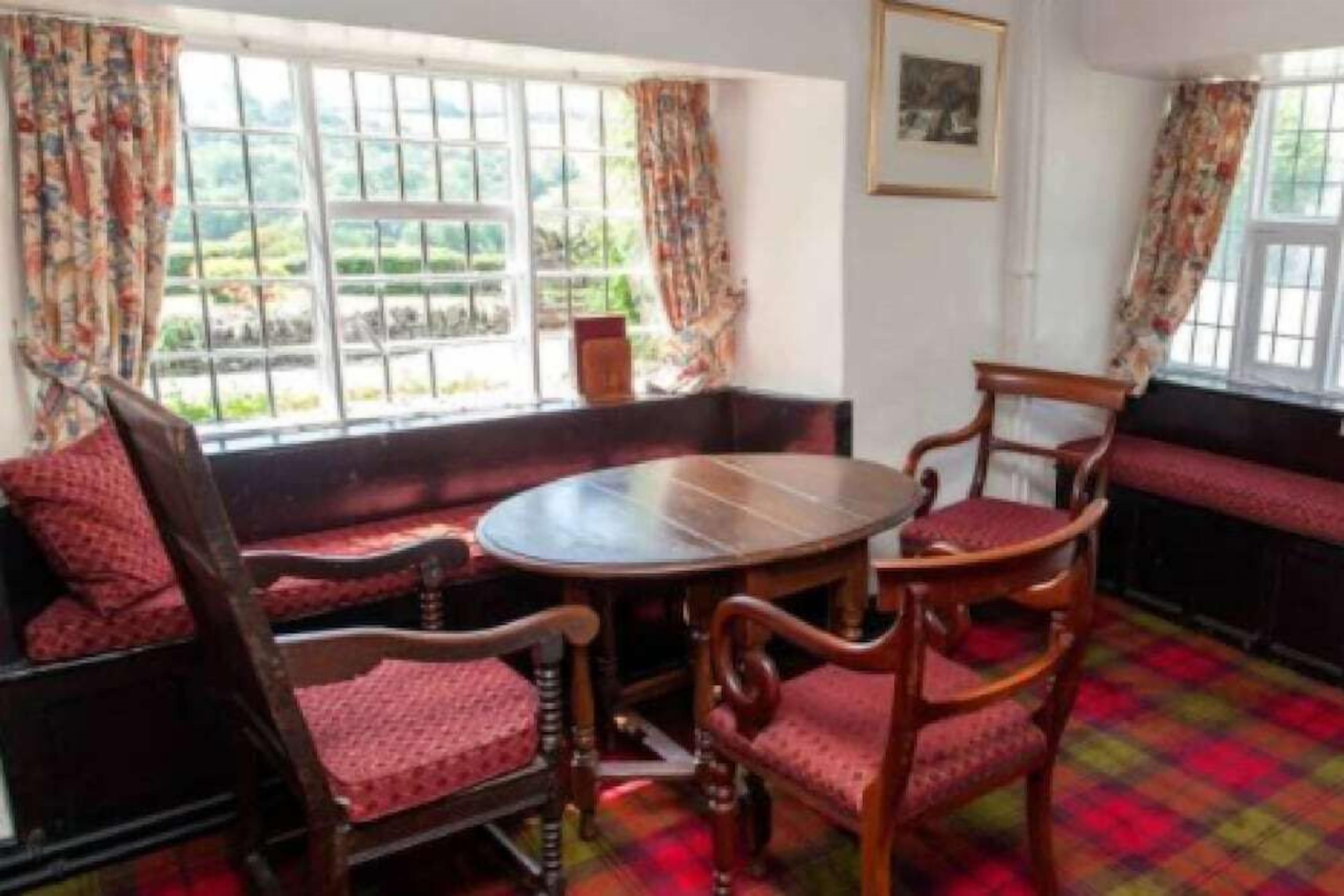 A room with a round wooden table, four wooden chairs with red cushions, a bench, and a window with a view of greenery.