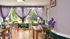 Un comedor con mesas y sillas de madera, cortinas moradas y un jarrón de flores sobre una mesa.
