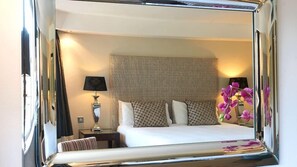 Een hotelkamer met een groot bed, een nachtkastje met een lamp en een spiegel die de kamer weerkaatst.
