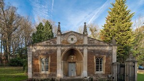 Ein kleines, historisches Gebäude mit spitzbogigem Eingang, einer Uhr über der Tür und einem kleinen Garten davor.