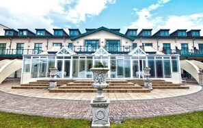 A building with a symmetrical facade, a central glass structure, and a paved courtyard with a fountain. - The Vale Golf Club (Pontyclun)