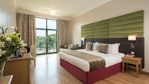 A hotel room with a large bed, a desk with a chair, a balcony with a view of greenery, and a chandelier.