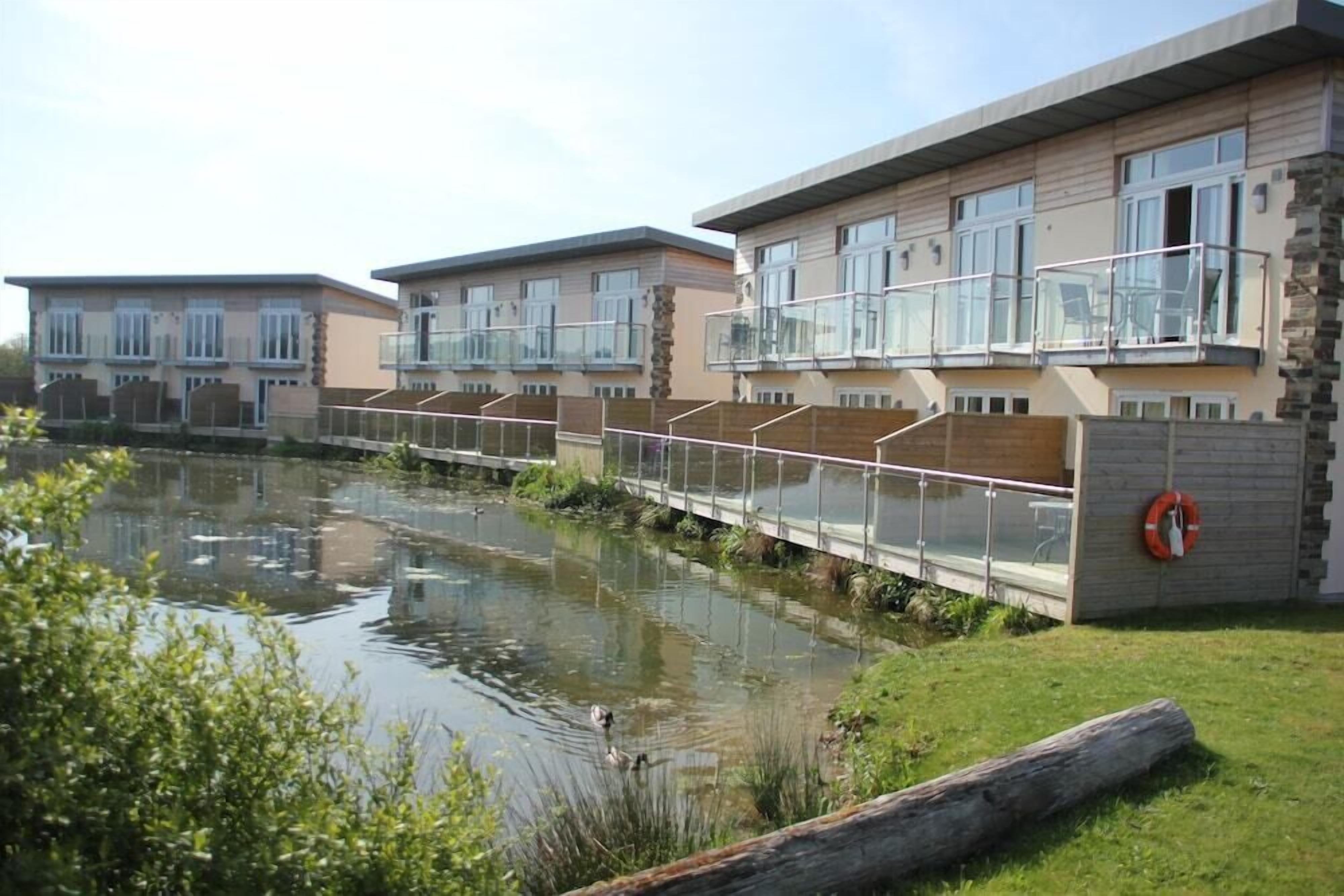 A modern residential building with a glass balcony, a small water body in front, and a grassy area.
