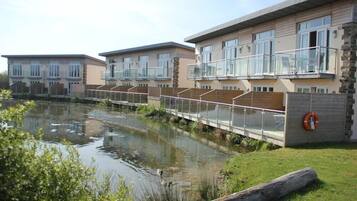 A modern residential building with a glass balcony, a small water body in front, and a grassy area.