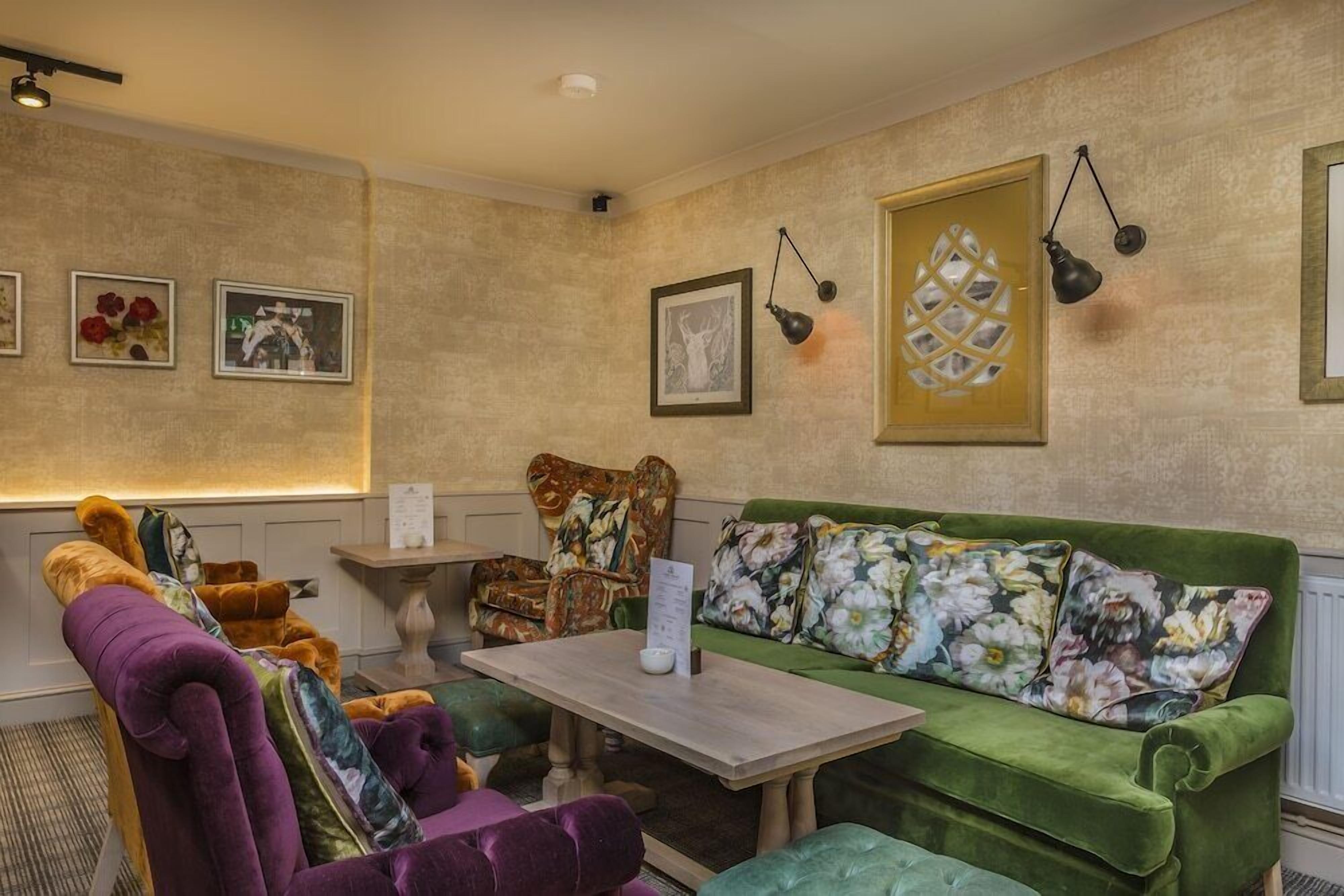 A cozy room with a green sofa, purple armchair, and a wooden table. There are framed pictures on the walls and a lamp hanging above the sofa.