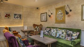 A cozy room with a green sofa, purple armchair, and a wooden table. There are framed pictures on the walls and a lamp hanging above the sofa.