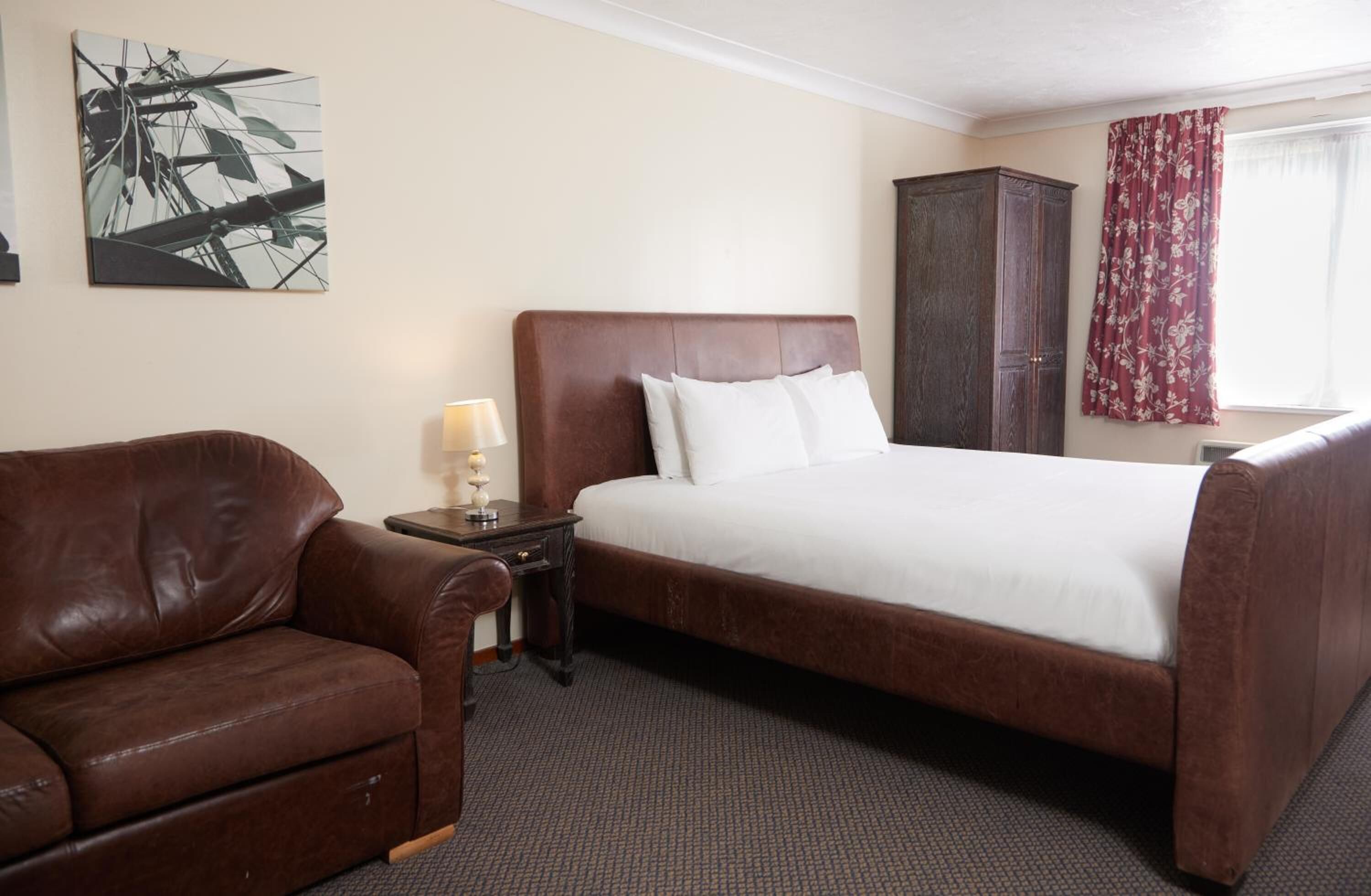 Een hotelkamer met een groot bed, een bruine leren bank, een klein tafeltje met een lamp, een kast en een raam met gordijnen.