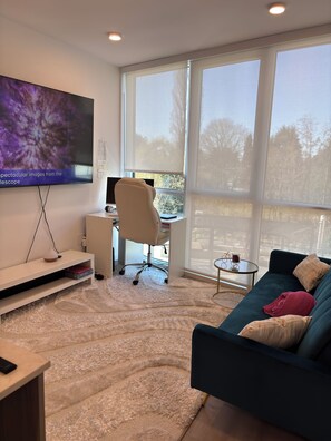 A modern living room with a large flat-screen TV, a desk with a computer, a comfortable sofa, and a view of trees through large windows. - Cozy 2-bedroom apartment with AC in peaceful Surrey (Surrey)