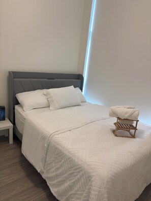 Cama bem arrumada com cabeceira cinza, roupa de cama branca e um pequeno banquinho de madeira com uma toalha em cima.