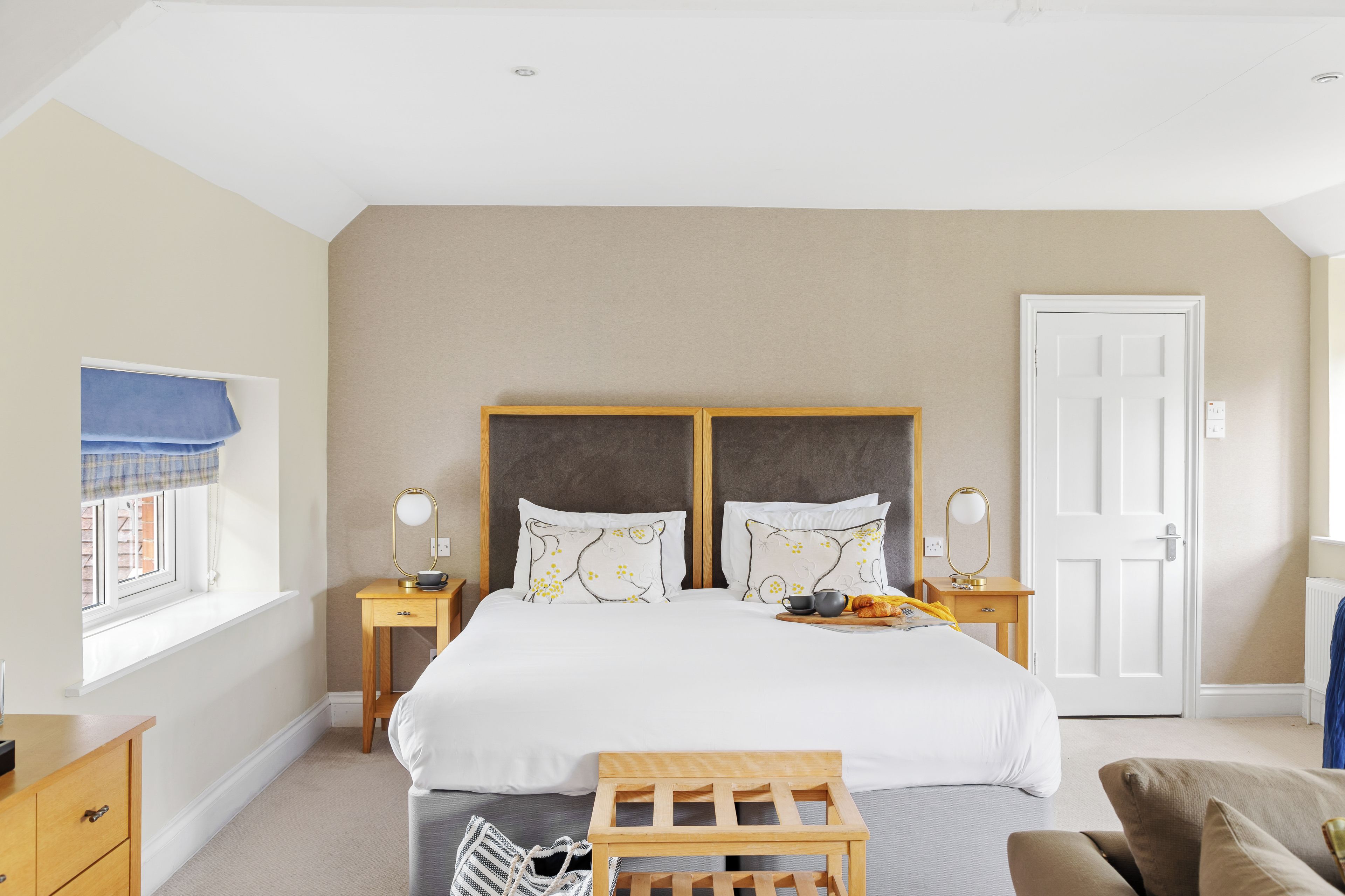 A hotel room with a large bed, two bedside tables, a window with a view, a door, and a small wooden bench.