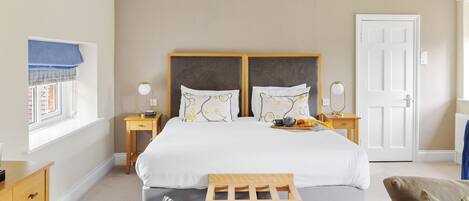 Habitación de hotel con una cama grande, dos mesitas de noche, una ventana con vistas, una puerta y un pequeño banco de madera.