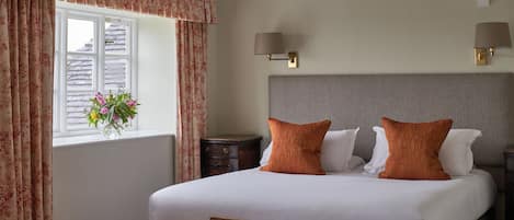 A bedroom with a large bed, two bedside lamps, a nightstand, a window with curtains, and a small wooden bench.