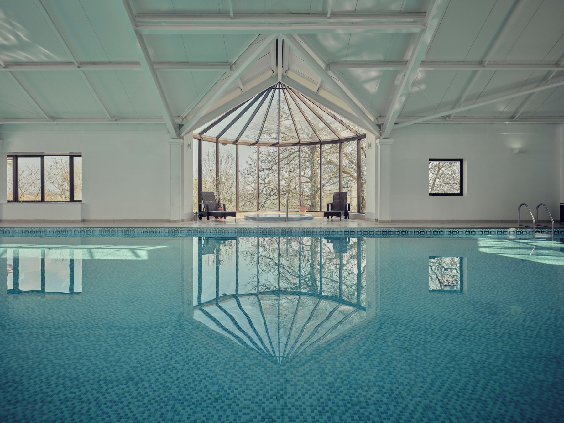 Una piscina interior con paredes de vidrio transparente, un techo alto y una zona central de observación con sillas.