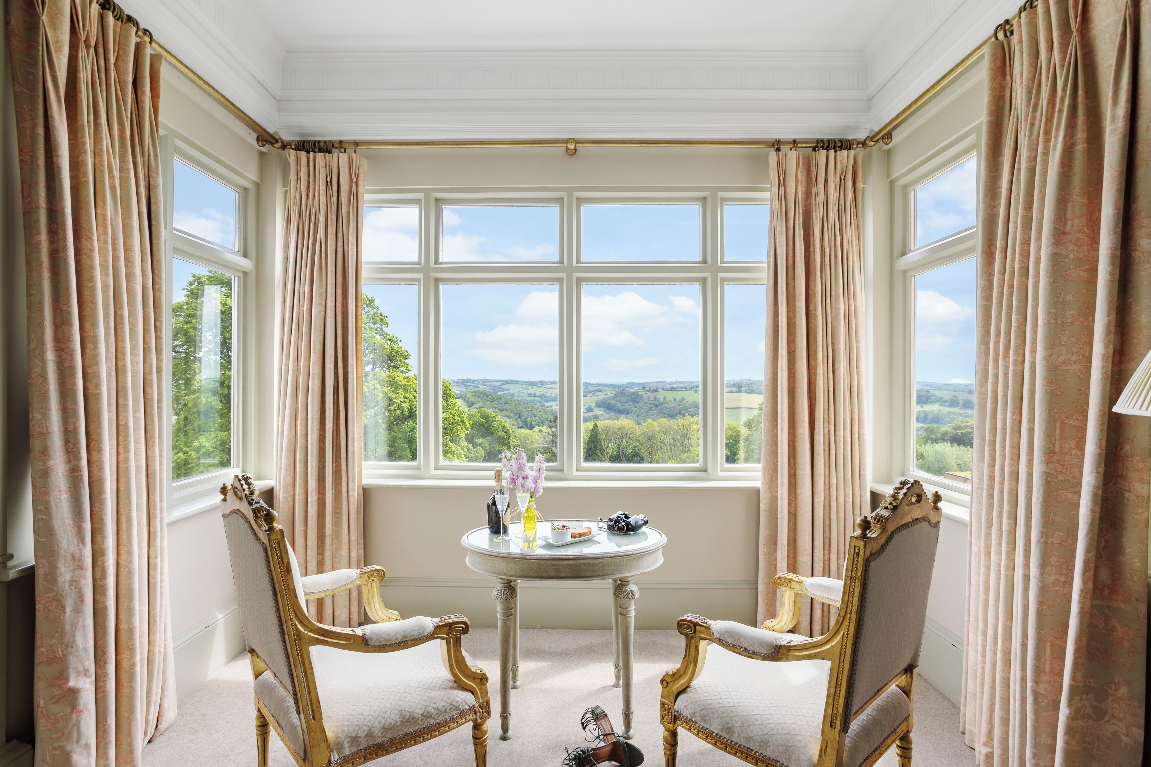 A room with a large window, two ornate chairs, and a small table with a bottle and flowers.