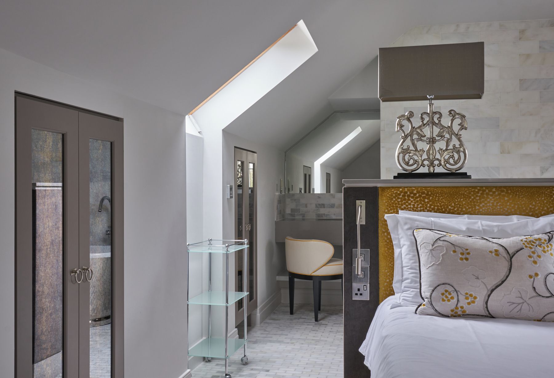 A modern bedroom with a glass shower, a chair, and a bed with patterned pillows.