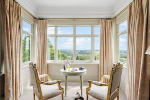 A room with two ornate chairs, a round table with a bottle and flowers, and a window with a view of greenery. - The Mole Resort (Umberleigh)
