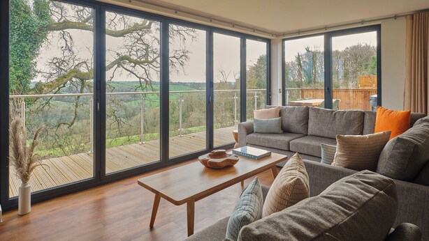 A modern living room with a large sliding glass door, a grey sofa, a wooden coffee table, and a view of a green landscape. - The Mole Resort (Umberleigh)