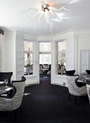 Une chambre d’hôtel dotée d’un lustre moderne, d’un coin repas avec chaises et tables, et donnant sur la chambre adjacente.