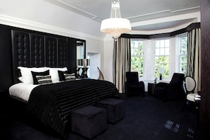 Une chambre d’hôtel avec un grand lit, un coin salon comprenant un fauteuil, une petite table et une grande fenêtre avec des rideaux.